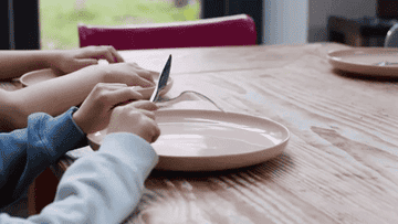A child plays with their cutlery over a pink dinner plate as they wait for food to be placed on the table.