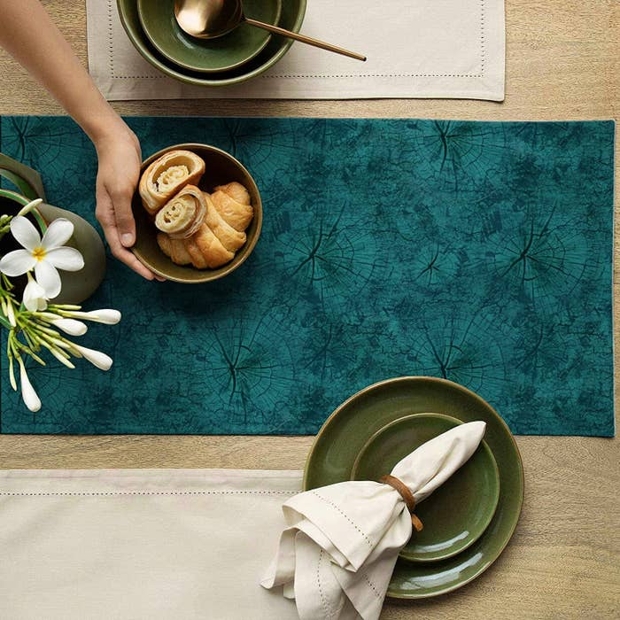 A teal blue suede table runner with green ceramic plates, a napkin, a bowl with baked items and a flower vase