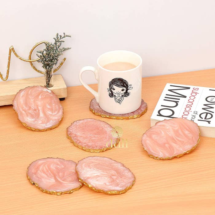 Six baby pink coasters with gold trimming arranged on a table with a book and a coffee mug