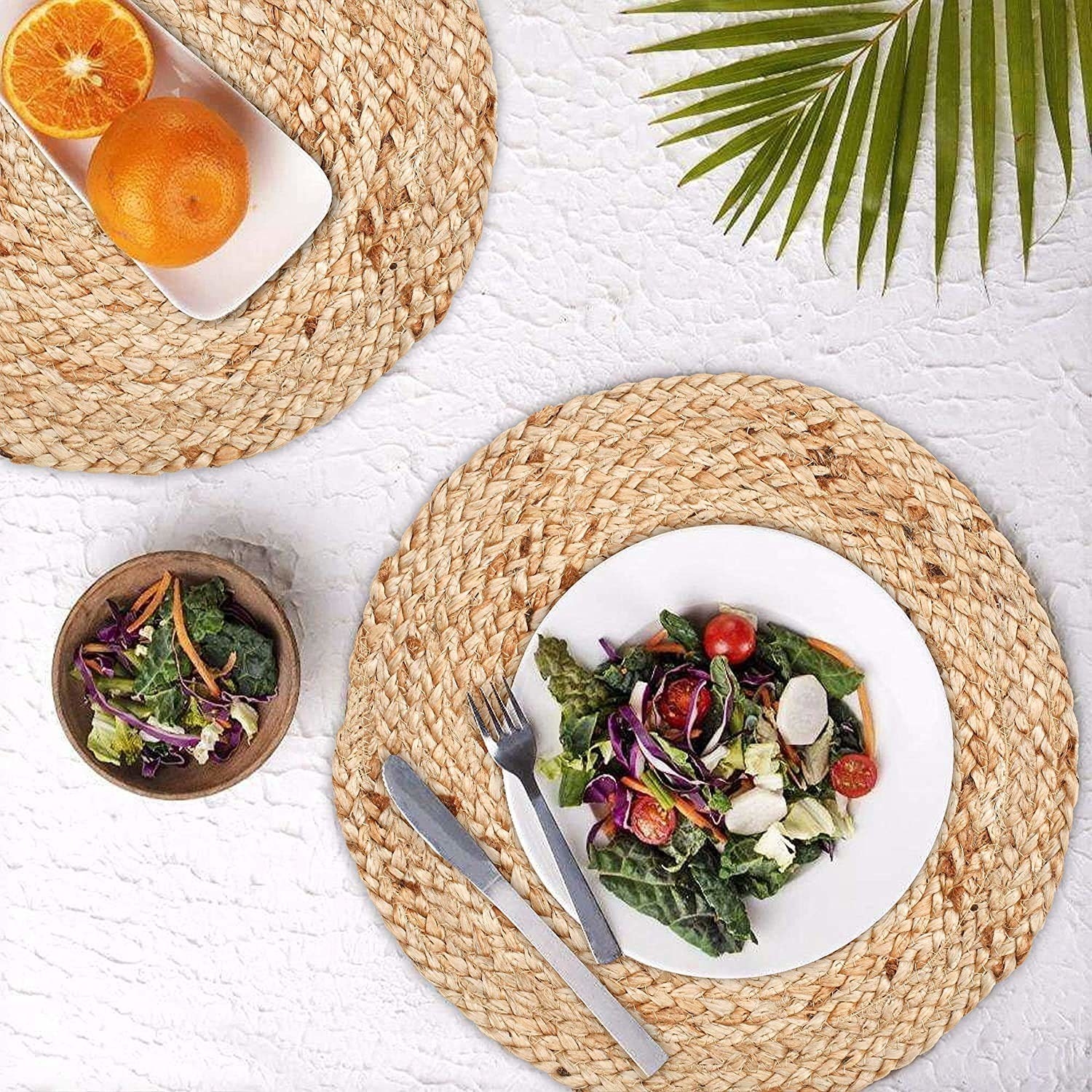 The round jute mats with two plates of food containing salad, a fork and knife and some oranges on another plate placed on a white background