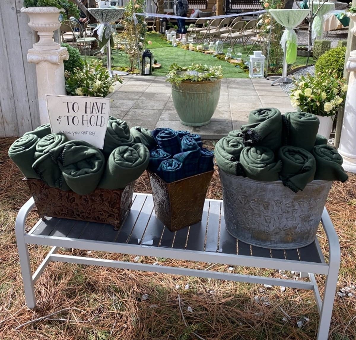 Dark green and navy blue blankets in baskets outside on a bench with a sign that says &quot;to have and to cold, in case you get cold&quot;