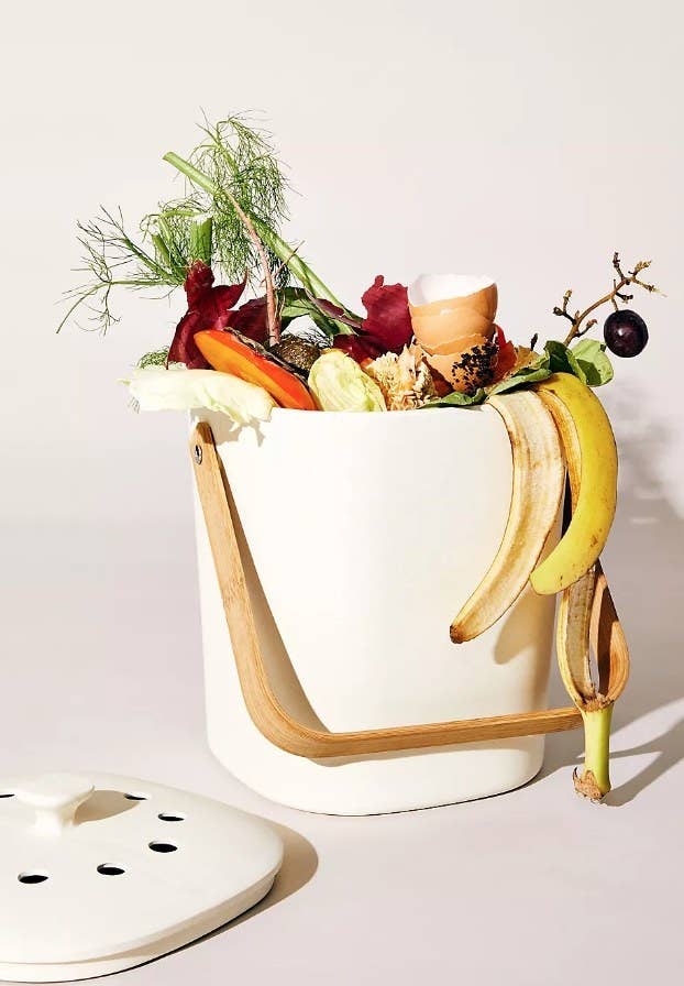 A cream, organic compost bin with a vented lid and bamboo handle filled with compost-friendly foods
