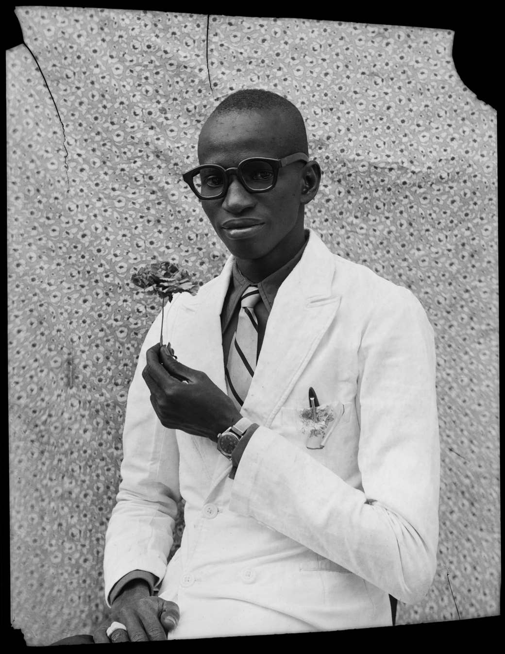 A man in a white suit holds a flower in front of a patterned background