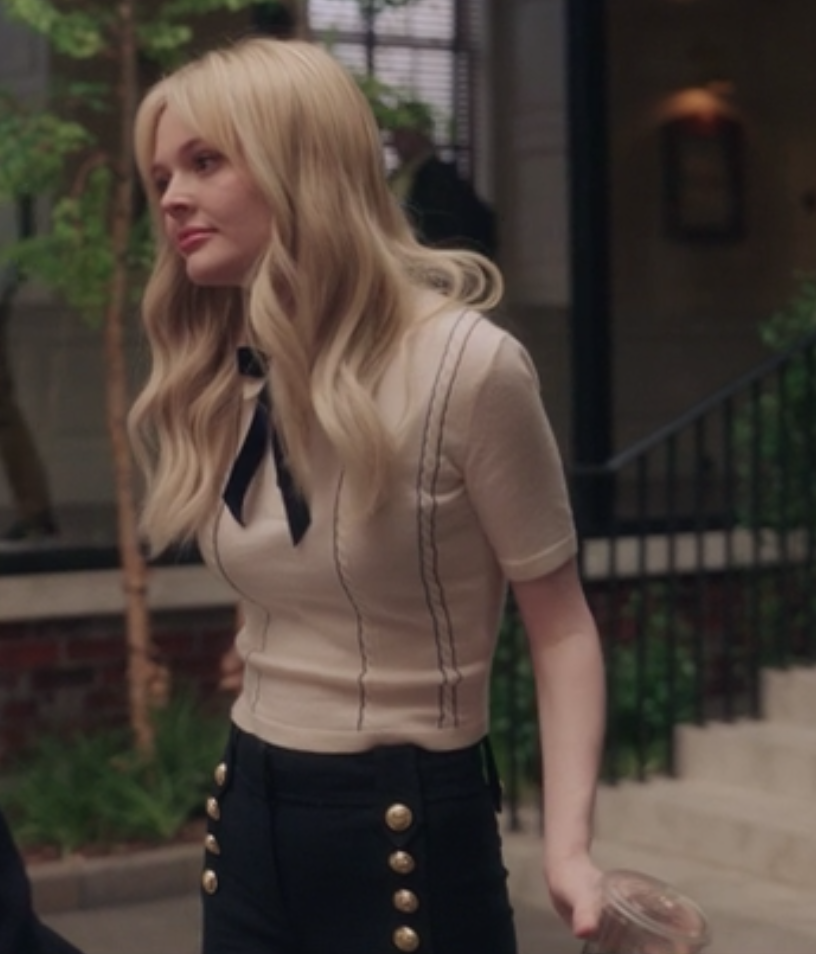 Audrey wears a knit sweater, oversize bow tie, and high-waisted pants with brass buttons on it