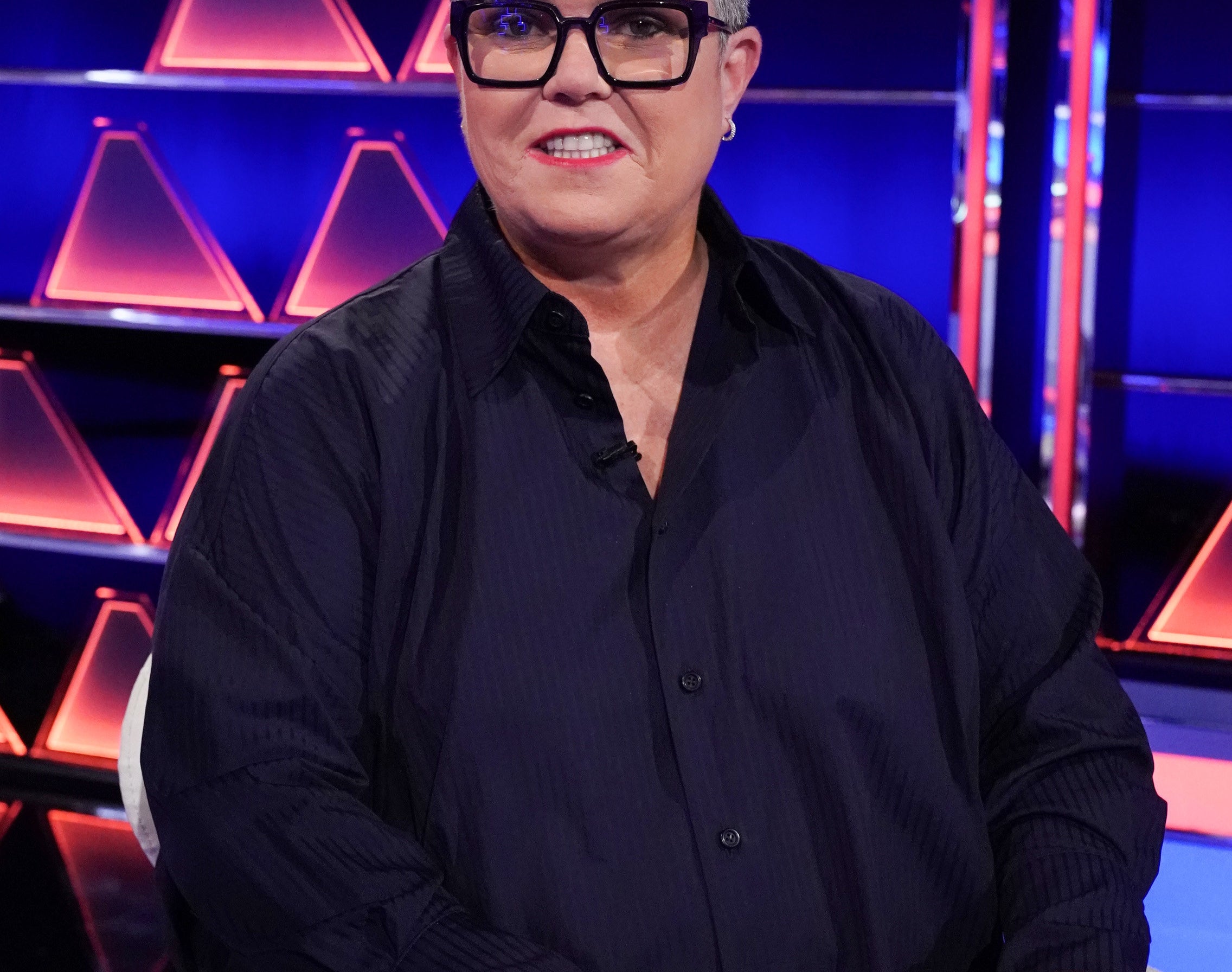 Rosie wears a black button down while smiling on the set of a game show