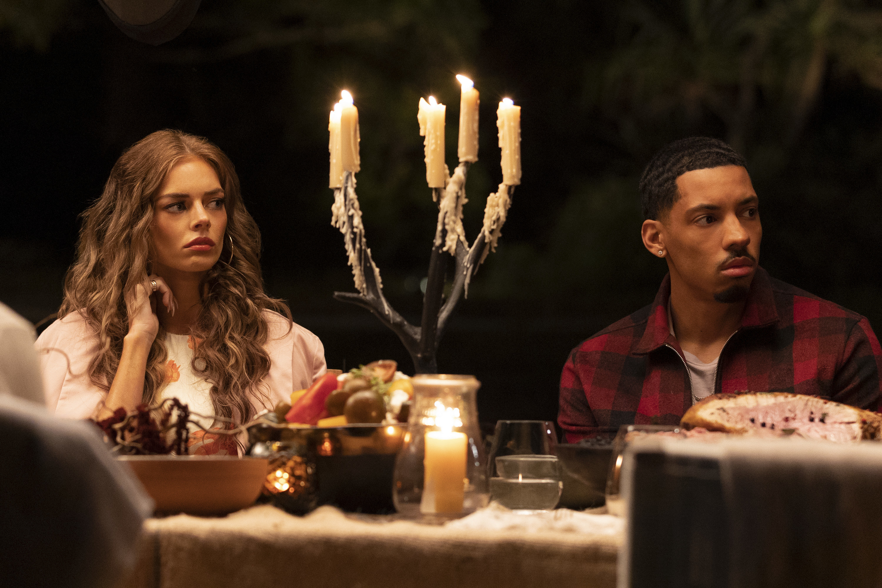 A close up of two characters sitting down at a candle-lit dinner table; they look worried