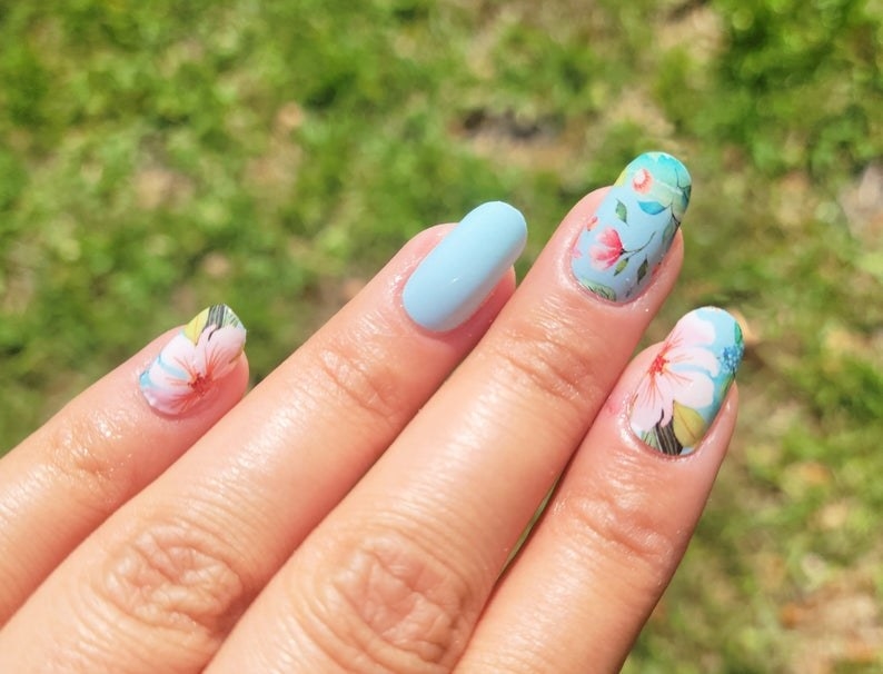 model&#x27;s hand showin three blue nails, two of the blue nails have flower decals on top while one is solid blue, and the pinky nail is one pink flower