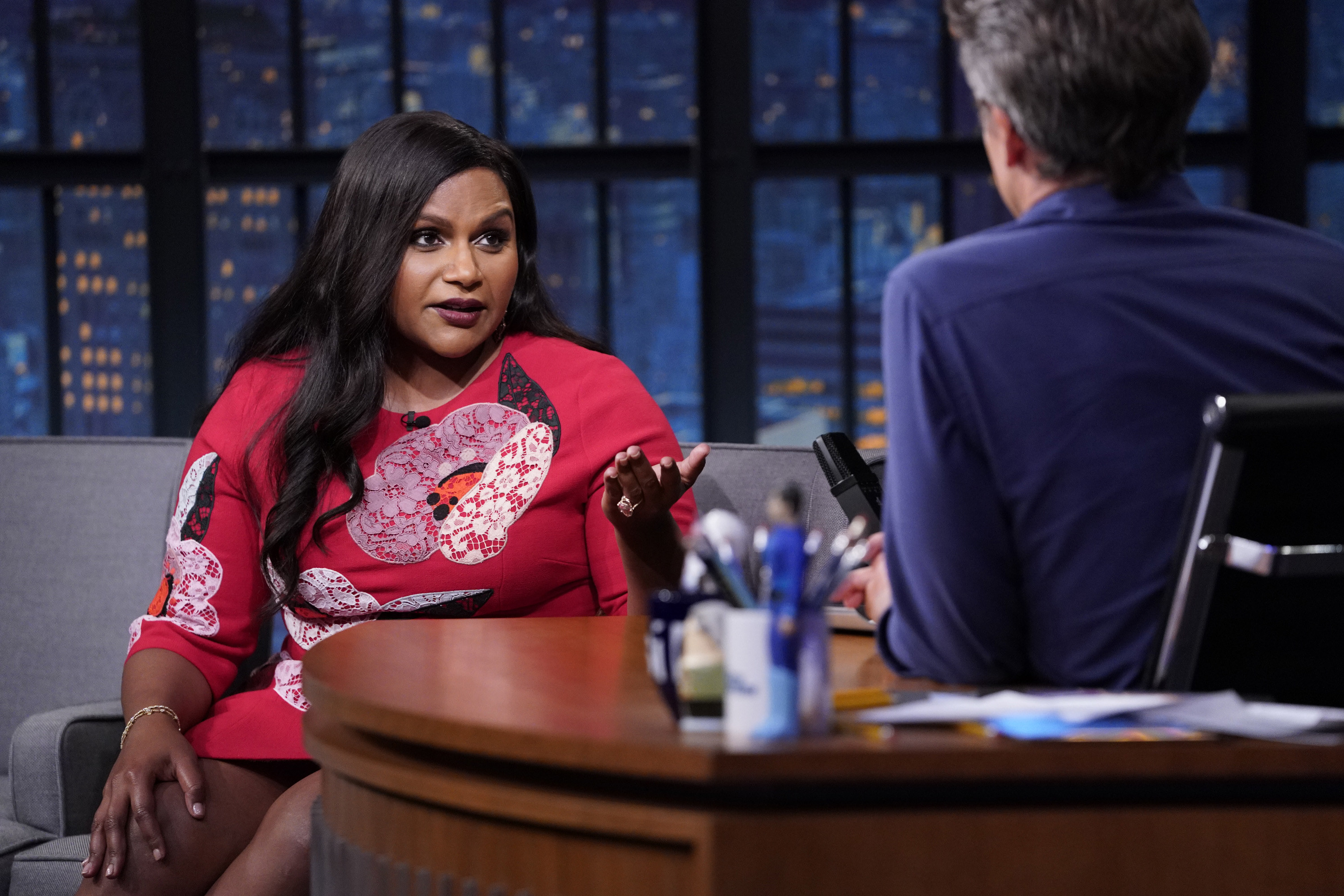 Photo of Mindy Kaling in a dark pink dress being interview by Seth Meyers