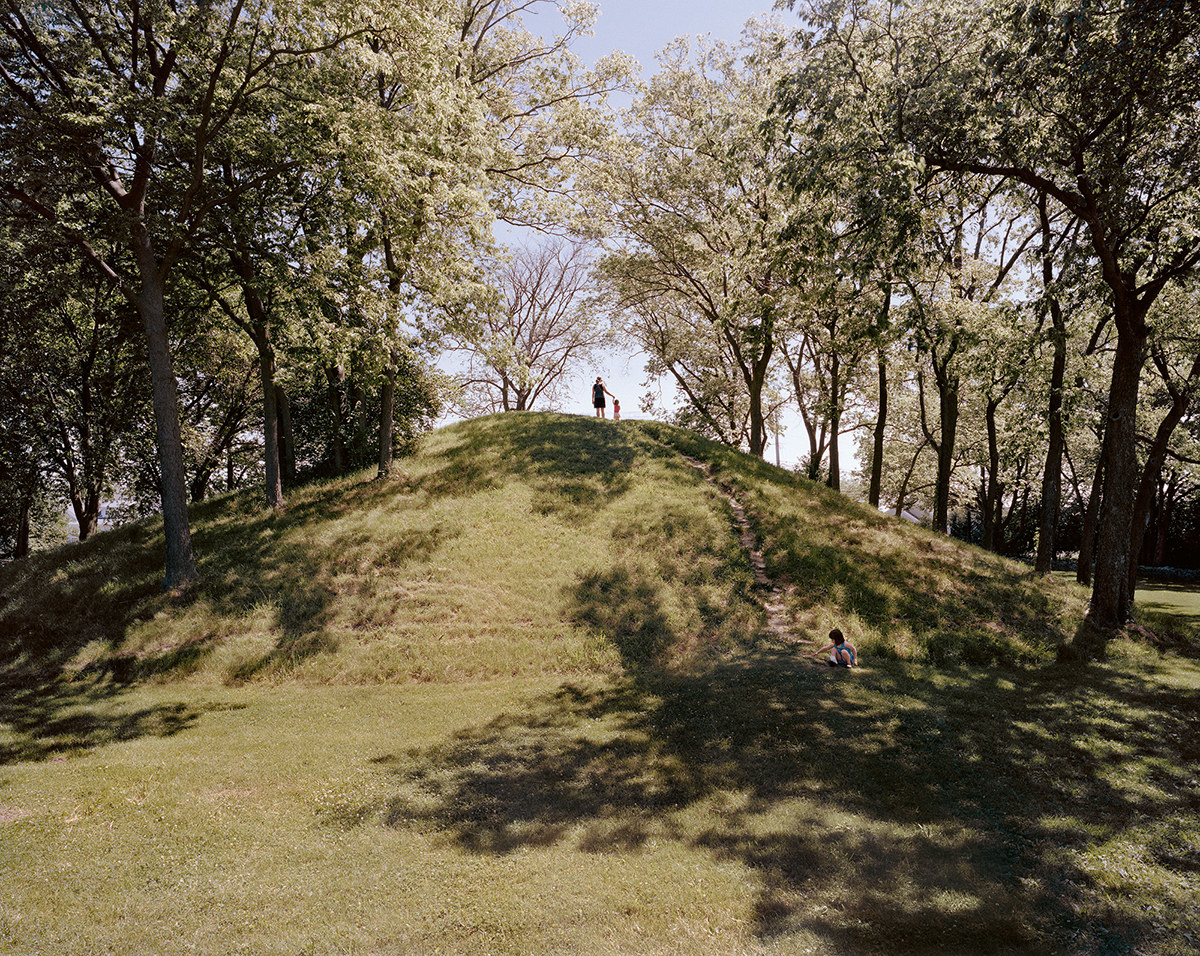 A woman and a child stand at the top of a big, grassy hill surrounded by trees, while another child crouches at the bottom