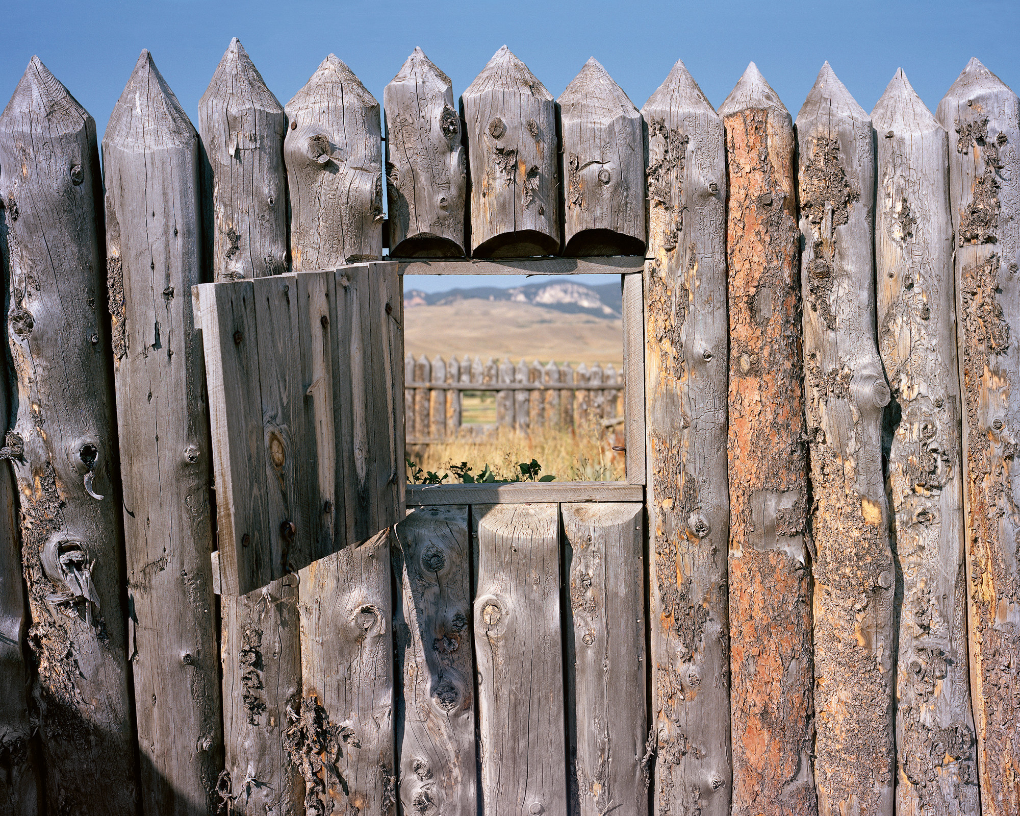 A window cut into walls made out of wood opens out into another row of wood walls and arid grassland