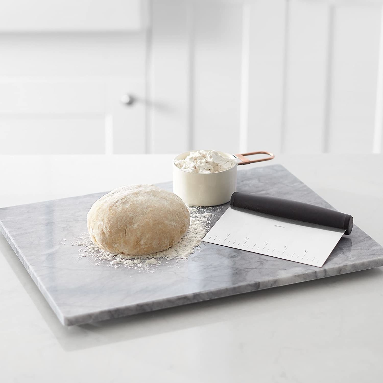 A stainless steel bench scraper kept beside a kneaded ball of dough and a cup of flour