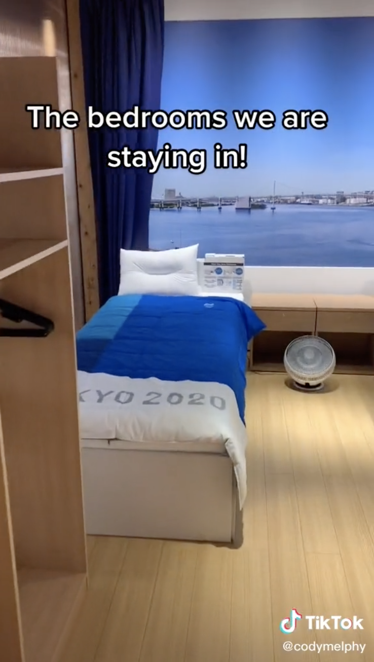 A twin-size bed with Tokyo 2020 written on the comforter and a fan on the floor
