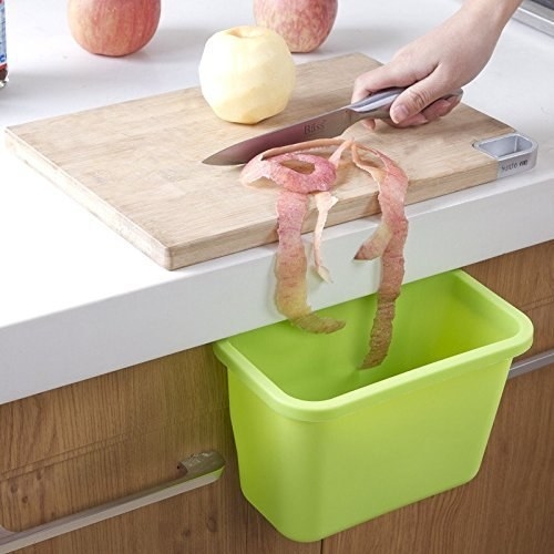 A person throwing an apple peel into a lime green hanging trash bin placed right below the counter