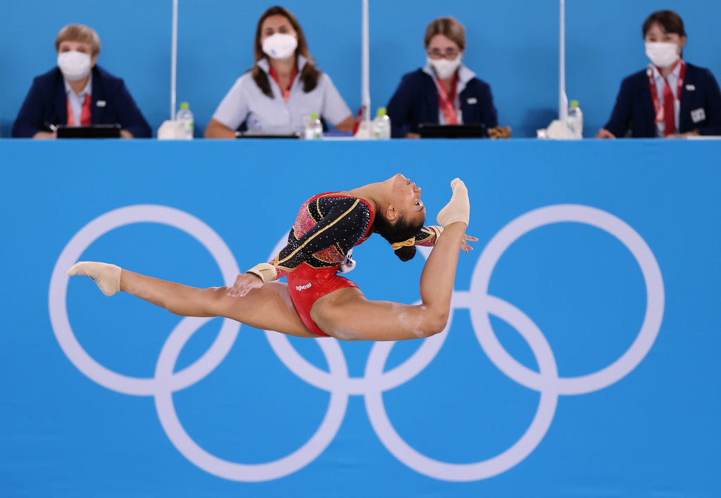 An airborne gymnast with one of their legs extended behind them and near their head