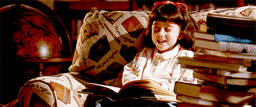 Matilda laughs hysterically while reading a book in a comfy armchair, surrounded by a pile of books