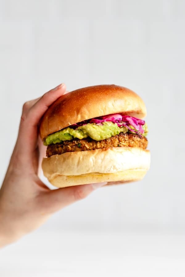 A hand holding a thick burger with layers of avocado and cabbage