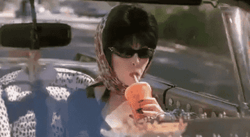 Elvira in sunglasses and a head scarf drinks from a soda cup while driving and pumps her fist in the air
