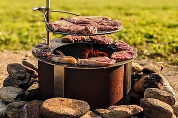 a copper colored circulate fire pit surrounded by stones with a grill piece attached to the side with meat on top of it