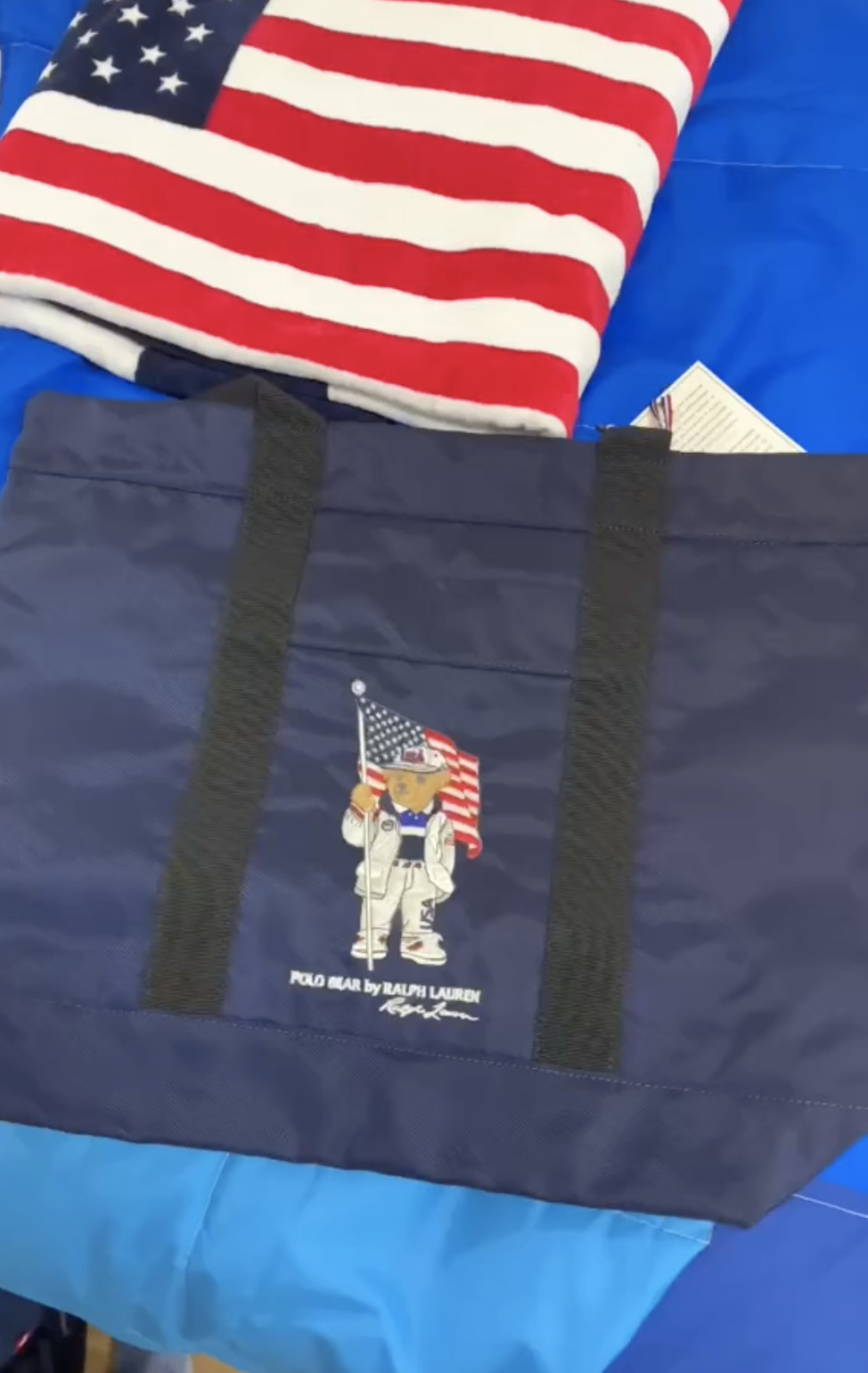 The tote has the Polo Bear by Ralph Lauren featuring an anthropomorphic bear wearing Ralph Lauren gear and holding a US flag