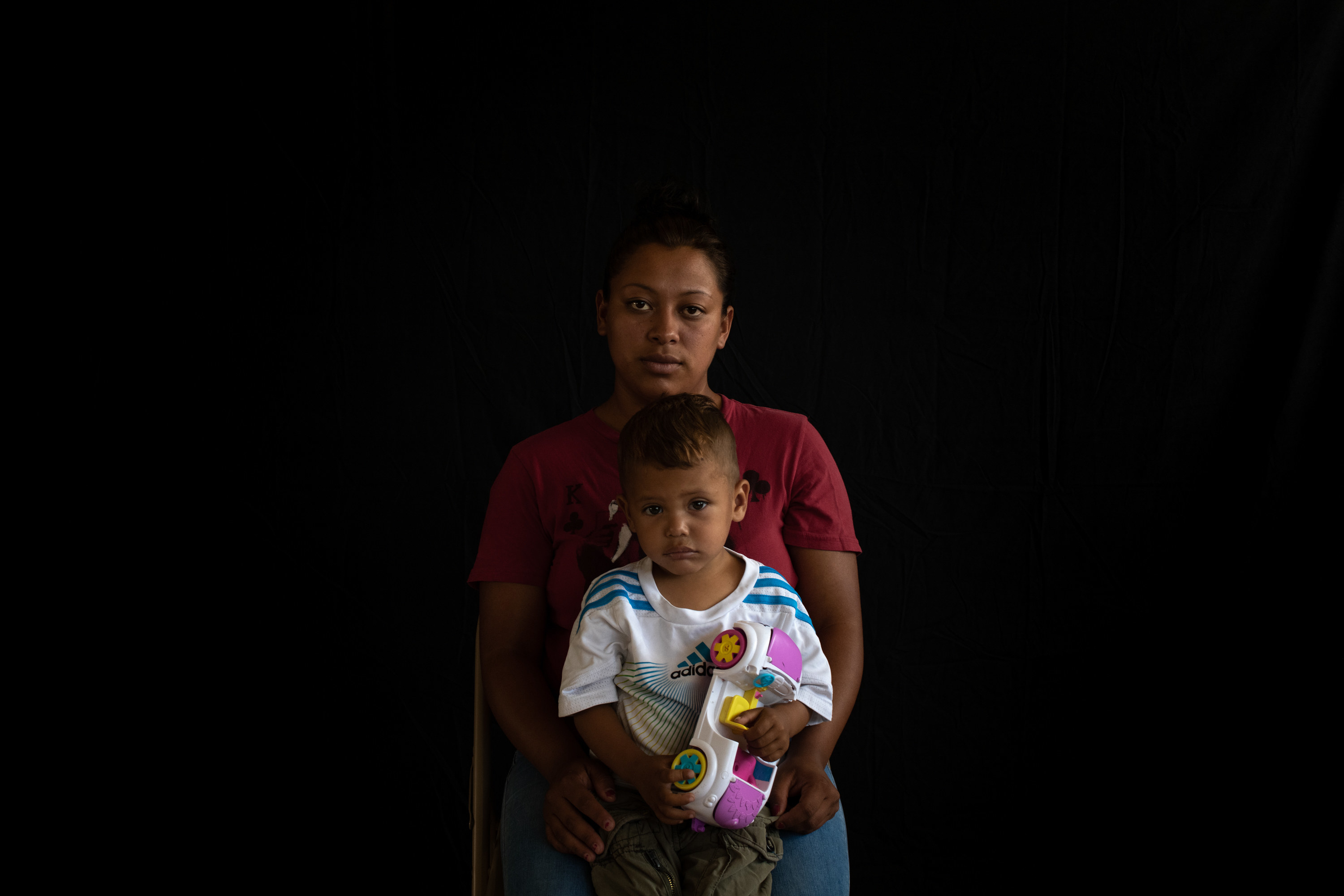 A woman sits in a folding chair, looking directly at the camera with her son in her lap, holding a toy car