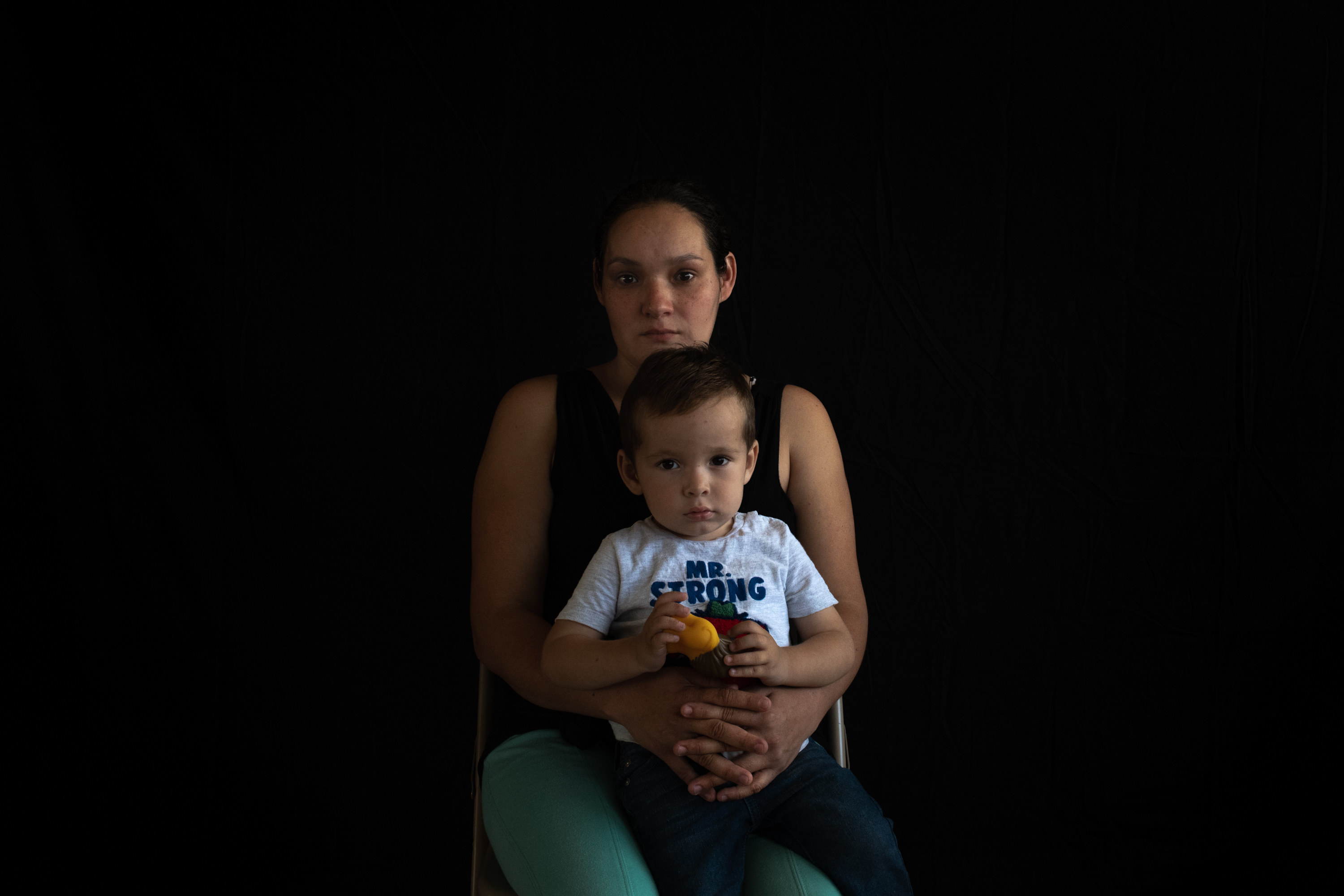 A young woman sits on a folding chair with a young boy on her lap. The boy is holding a toy lion and wearing a shirt that says &quot;Mister Strong&quot; 