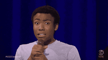 GIF of Donald Glover onstage cringing and pull his T-shirt neck