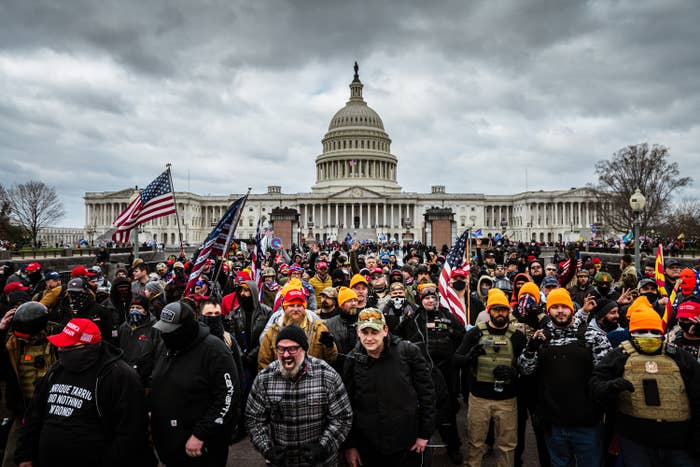 Hundreds of people, apparently mostly men, stand in front of the US Capitol with flags and some in tactical gear