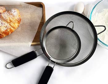 Two strainers on top of each other, laying beside flour and croissants 