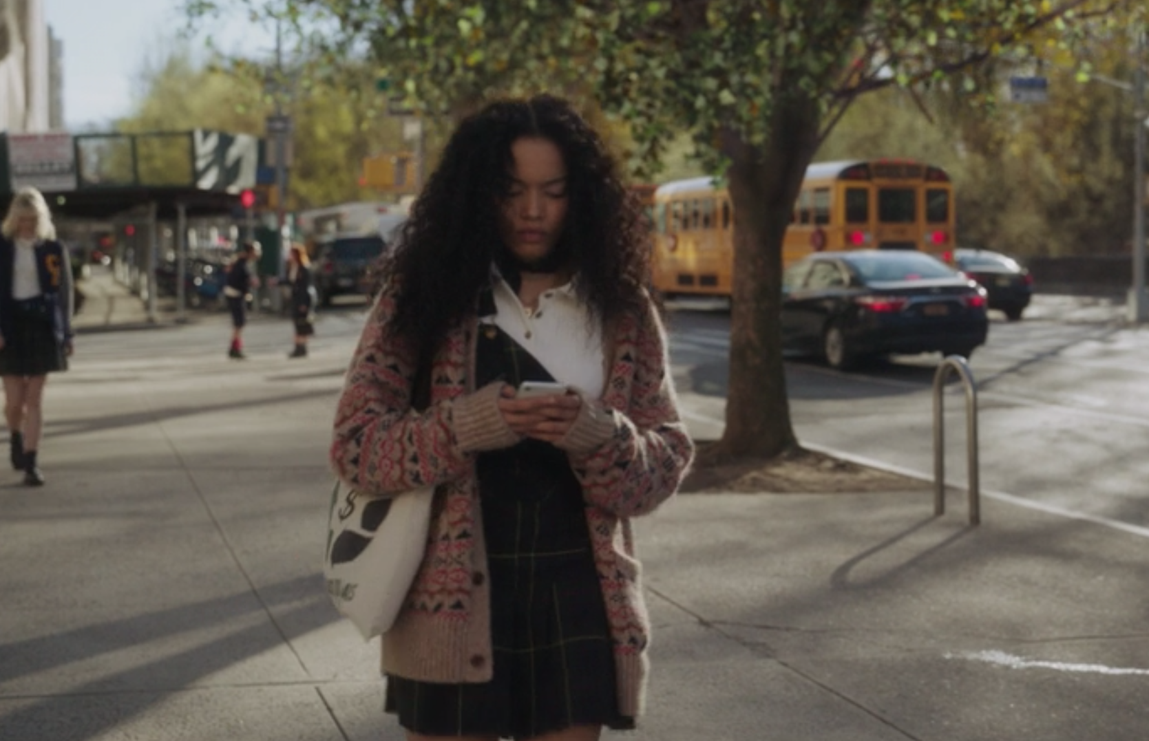 Zoya wears a plaid uniform jumper skirt with one strap attached to the top button, over a T-shirt with a patterned cardigan on top