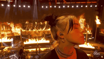 GIF of Billie looking at the camera with the set behind her aflame