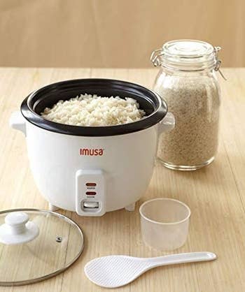 product shot of the cooker, lid, spoon, measuring tool, and uncooked rice in a jar 
