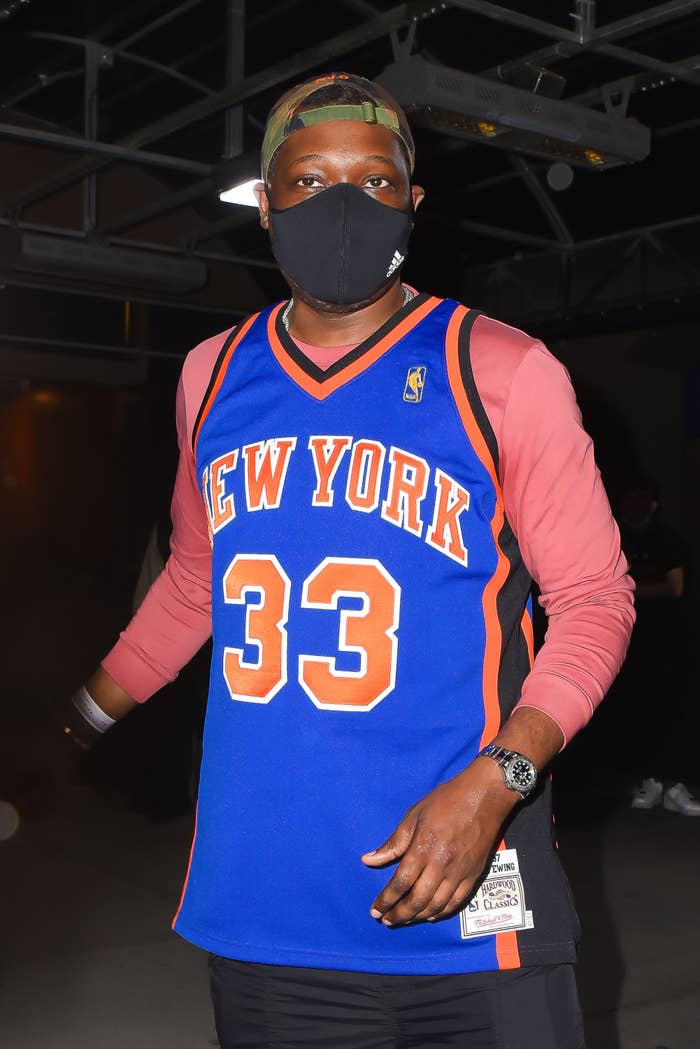 Michael Che walking outdoors in a New York Knicks jersey over a long-sleeved shirt, a bucket hat, and a face mask