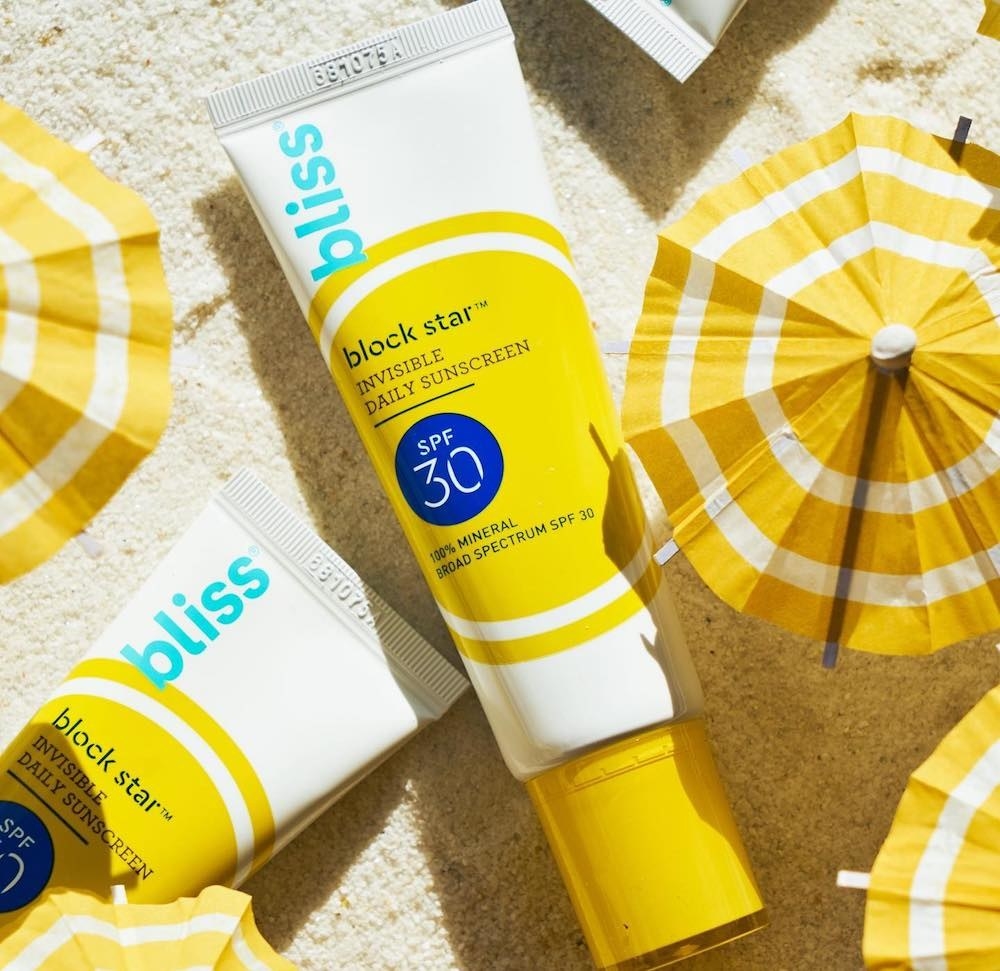 Bottles of Bliss Block Star mineral sunscreen on sand next to umbrellas