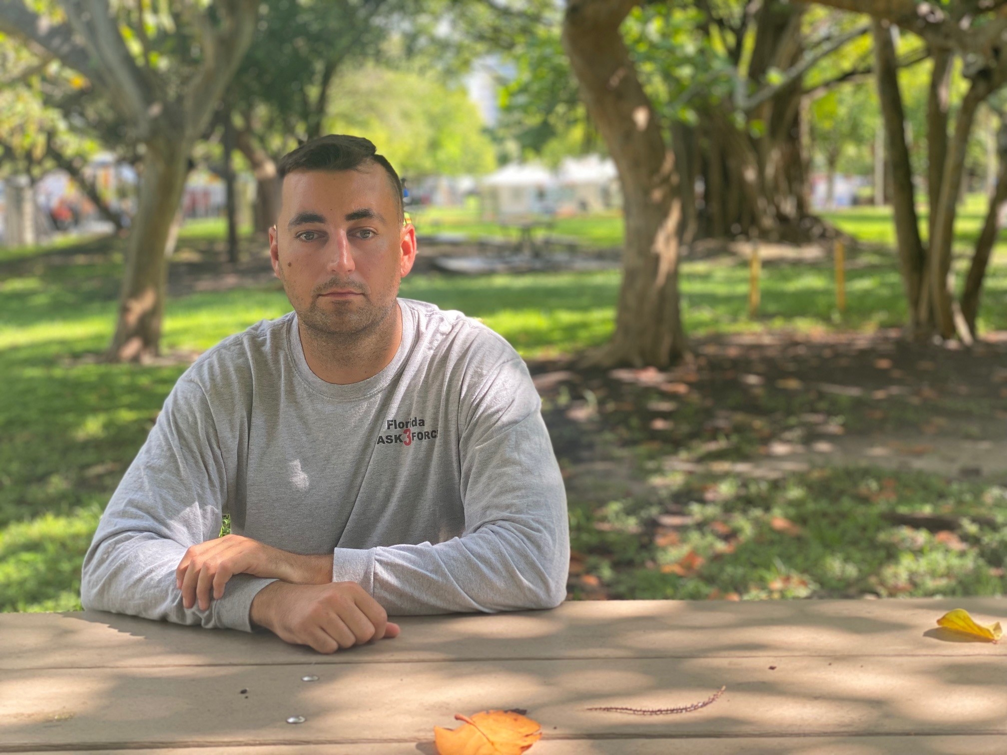 A somber man sits outdoors with his arms resting on a table at the elbows