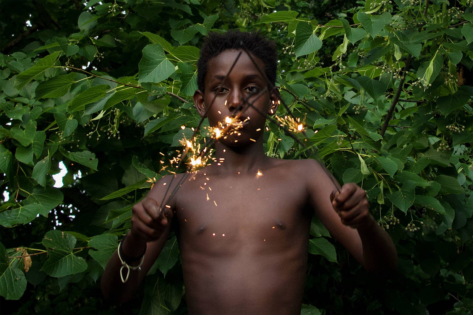A shirtless child standing in front of a bush holds up lit-up sparklers in both hands