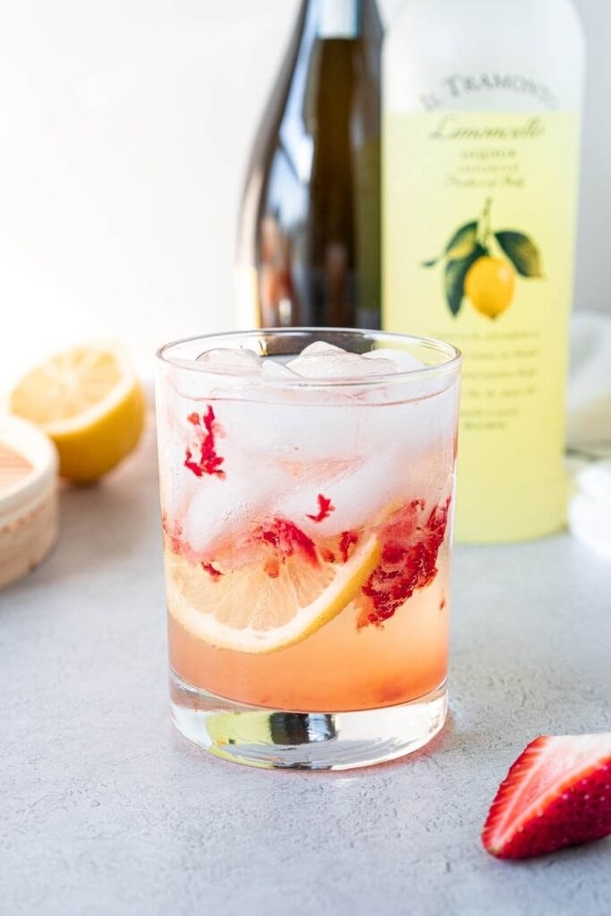 Pink cocktail in a glass with ice, lemon, and muddled strawberries. 