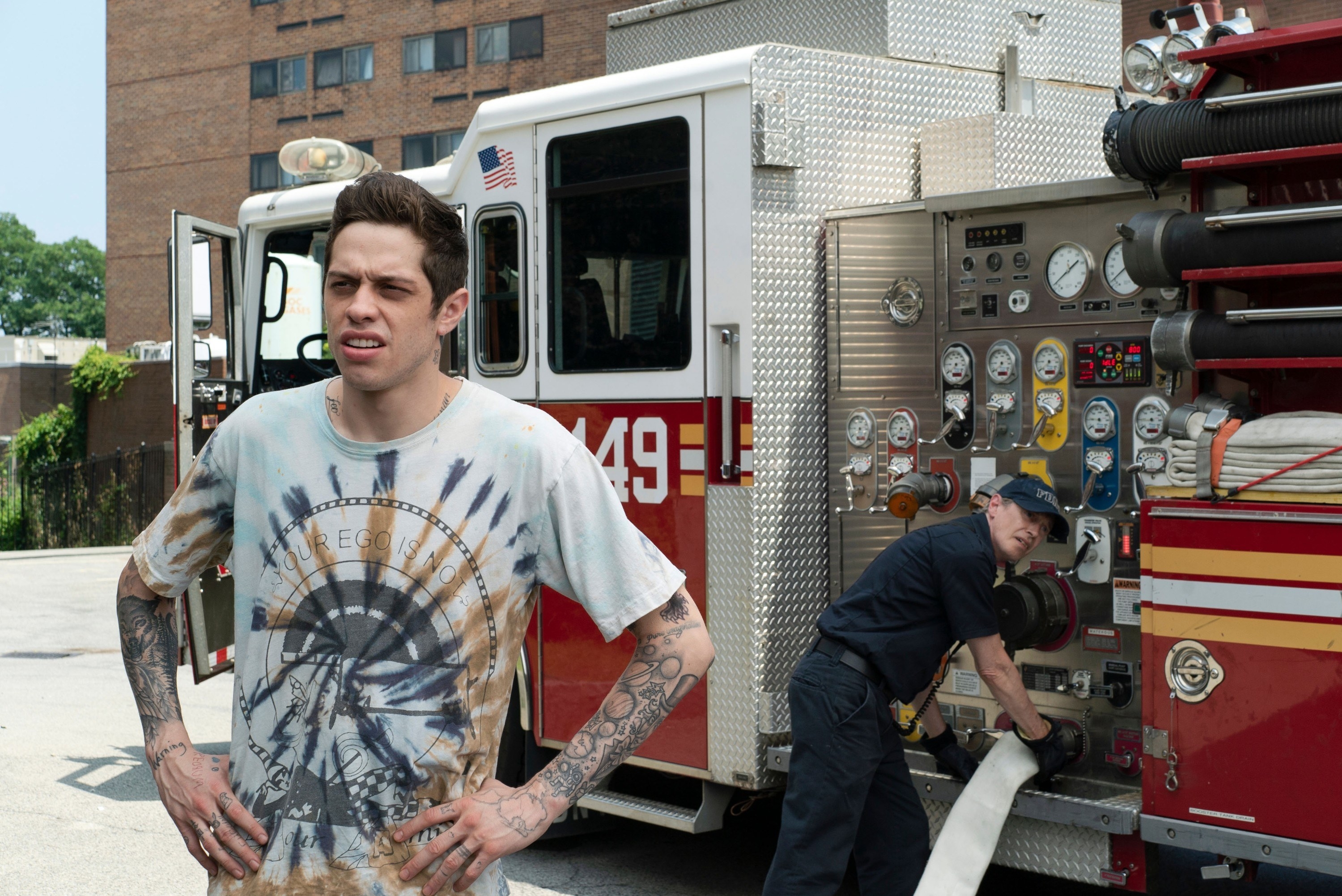 Pete Davidson poses in front of a firetruck (being worked on by Steve Buscemi) on set of &quot;The King of Staten Island&quot;