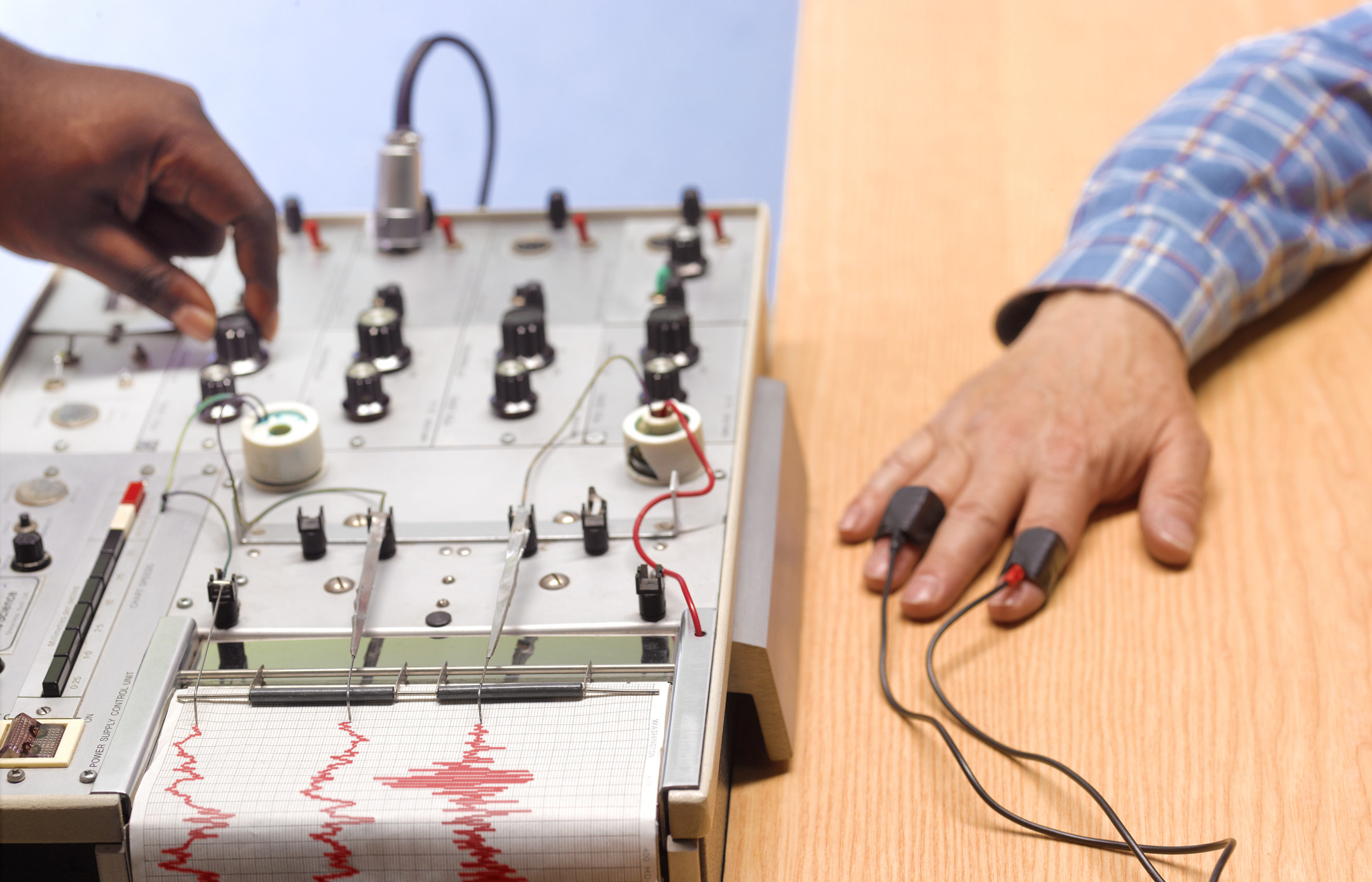 A polygraph test with a hand on one knob across from a man&#x27;s hand on the table connected by sensors on index and ring fingers