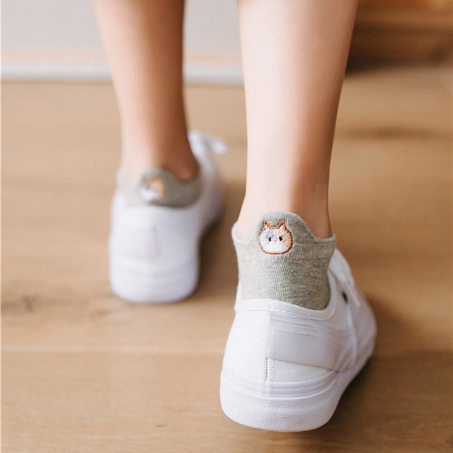 Model is wearing white tennis shoes with grey socks that have embroidered cats on the back