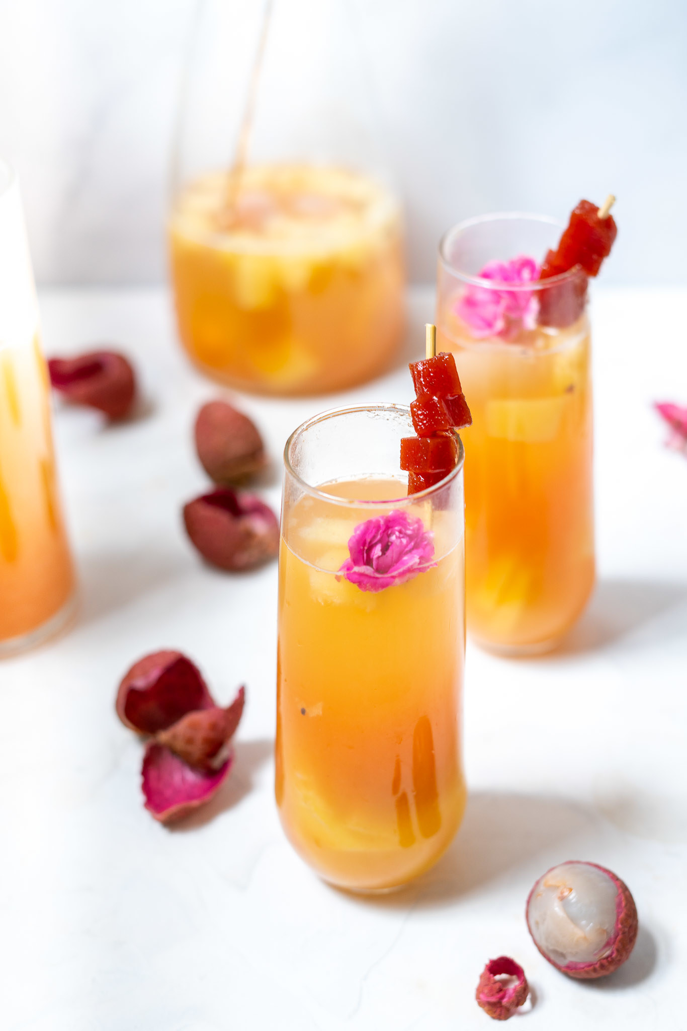 An orange colored cocktail in a stemless cocktail glass garnished with flowers and guava.