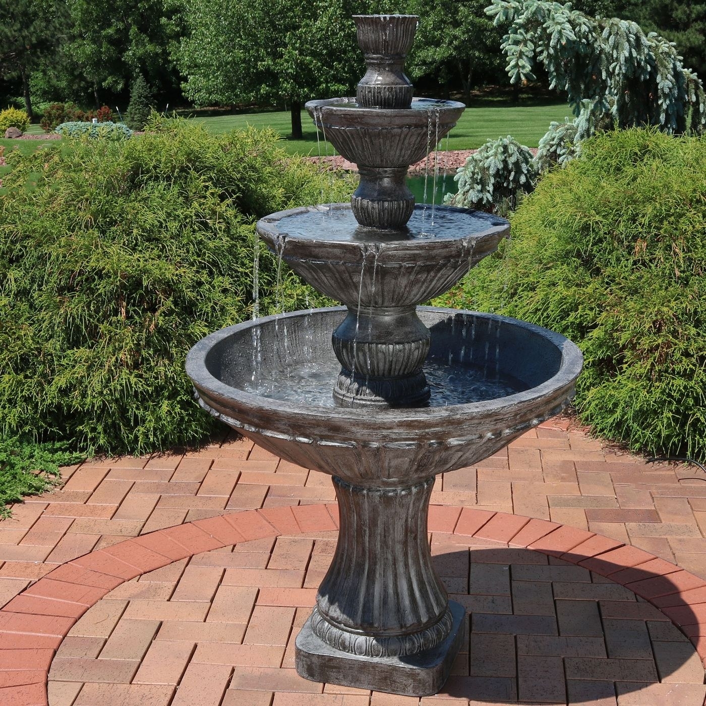 The three tiered fountain is grey and streams of water against a brick and green outdoor area