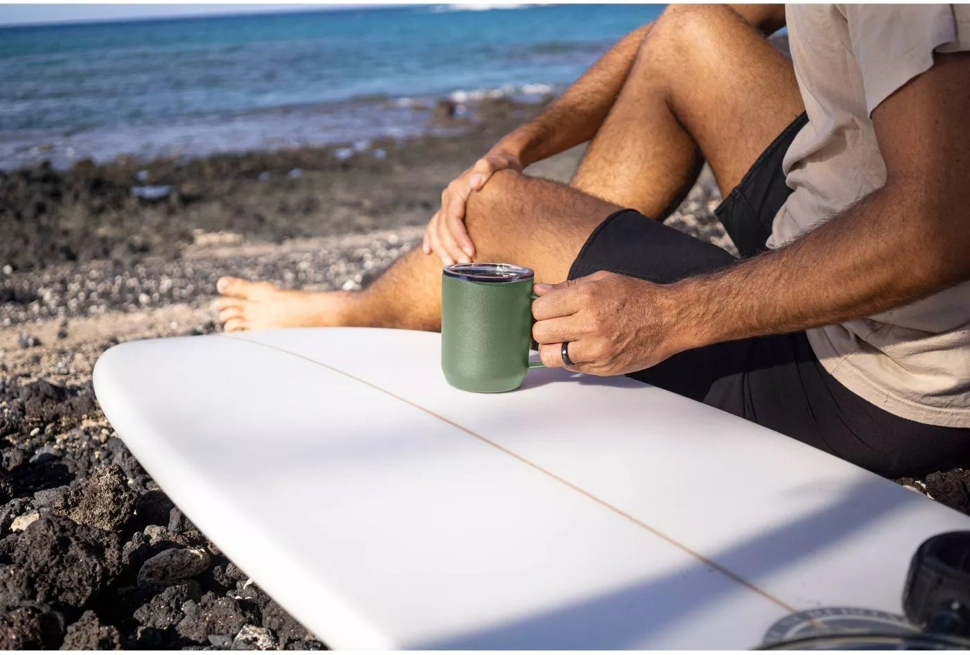 The green mug is in an adult's hand and is resting on a surfboard with a body of water in the background