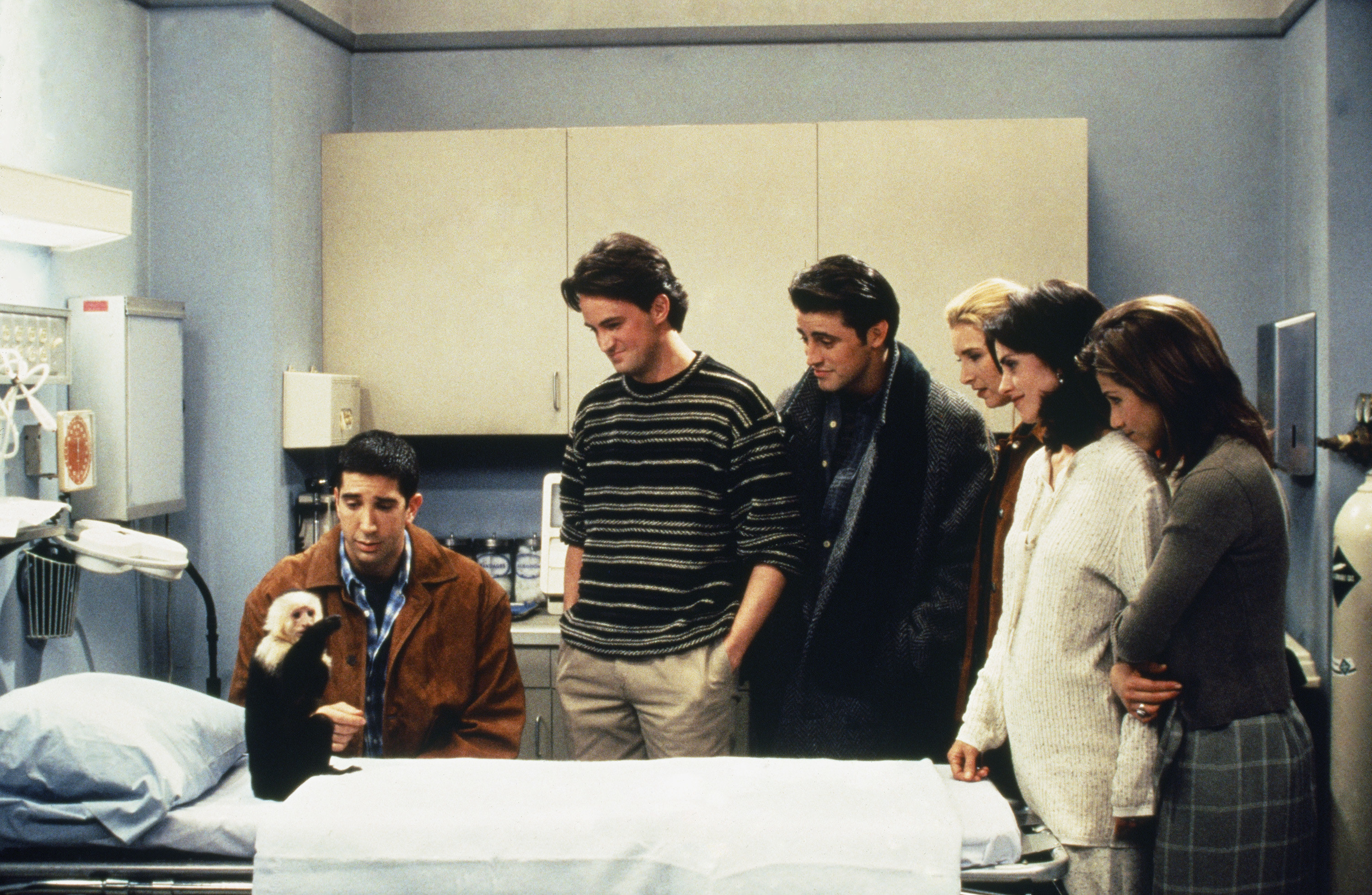 Ross and the rest of the cast look at Marcel while the monkey sits on a doctor&#x27;s table