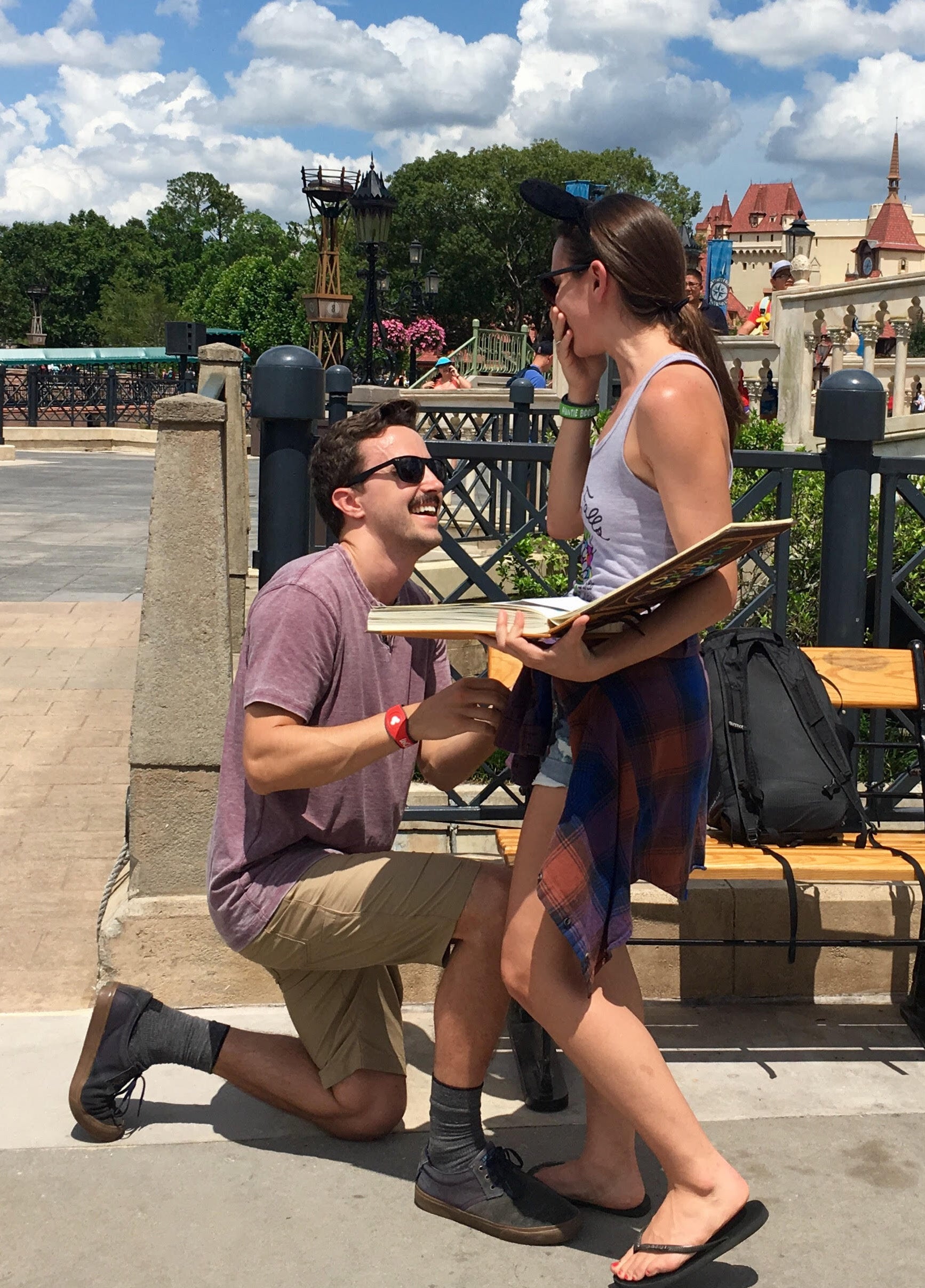 A man in sunglasses kneels in front of a woman with her hand over her mouth in surprise