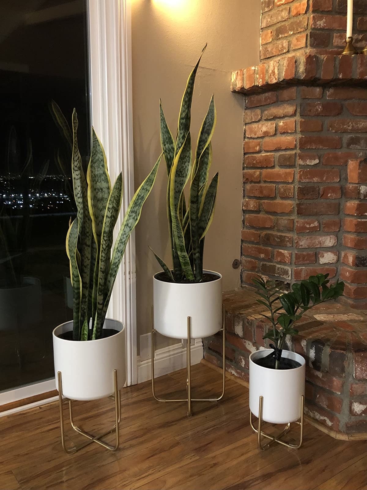 Reviewer&#x27;s set of three white planters with gold stands sit in the corner of a living room next to a fireplace