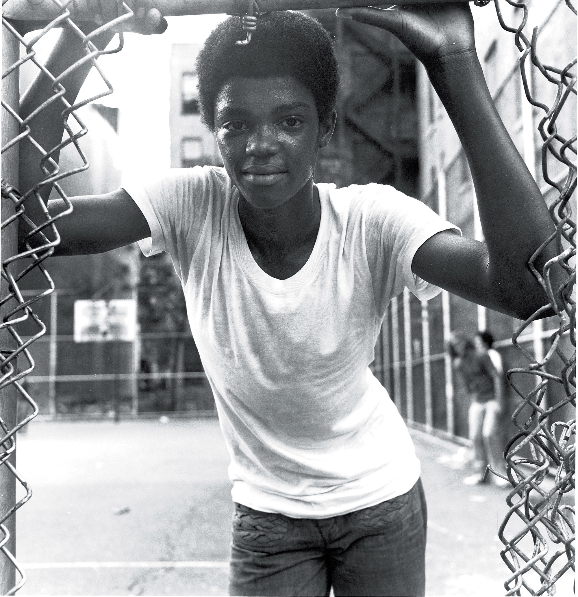 A young Black person smiles and stands by the side of a basketball court while looking through a gap in a chain-link fence