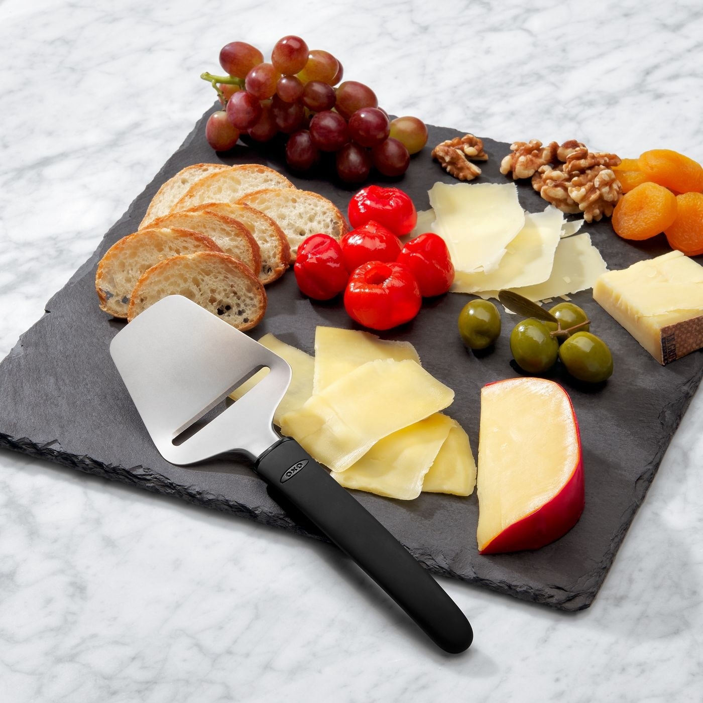 a charcuterie board with cheese, olives, bread, and fruits next to a cheese slicer