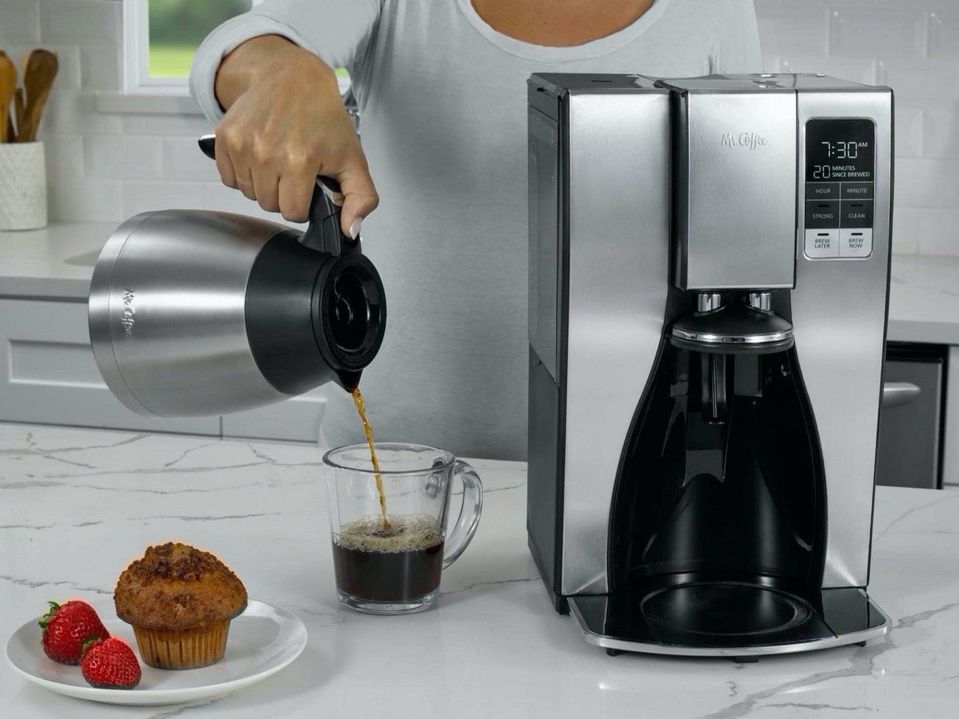 a woman pouring coffee into a coffee mug next to a coffee maker, a carafe, and a plate with a muffin and strawberries