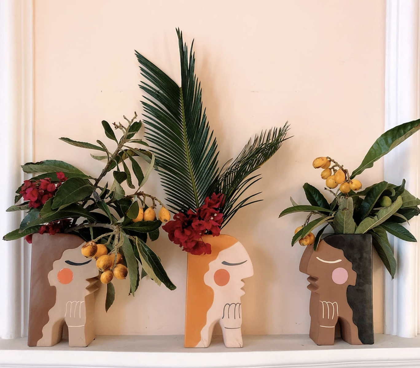 Three vases with different skin tones and hair colors display various types of leaves and fruit branches