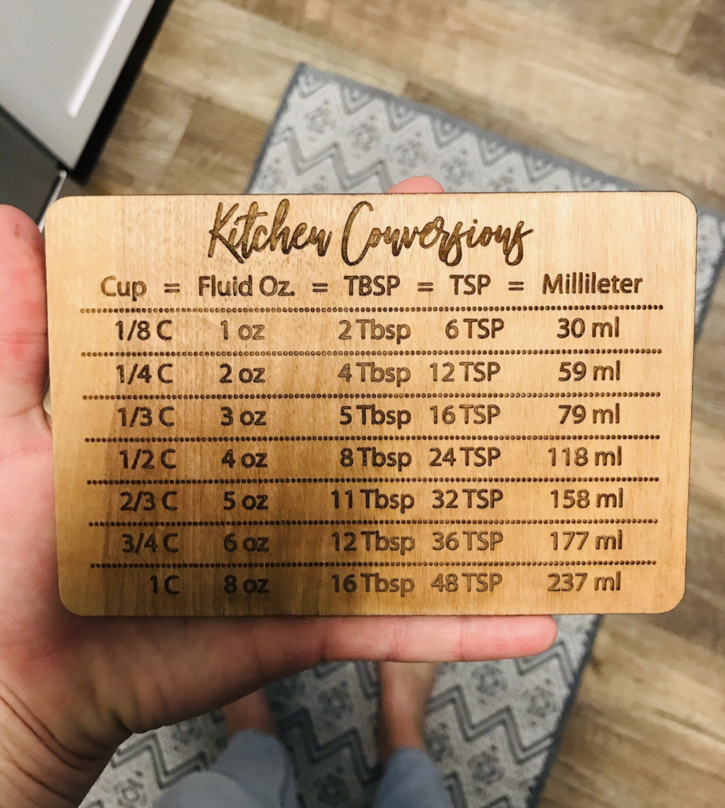 Person holding a wooden kitchen conversion chart with measurements for cups, fluid ounces, tablespoons, teaspoons, and milliliters